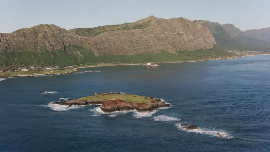 An aerial of the ocean coast up against a mountain range as waves crash against an island formation off the coast