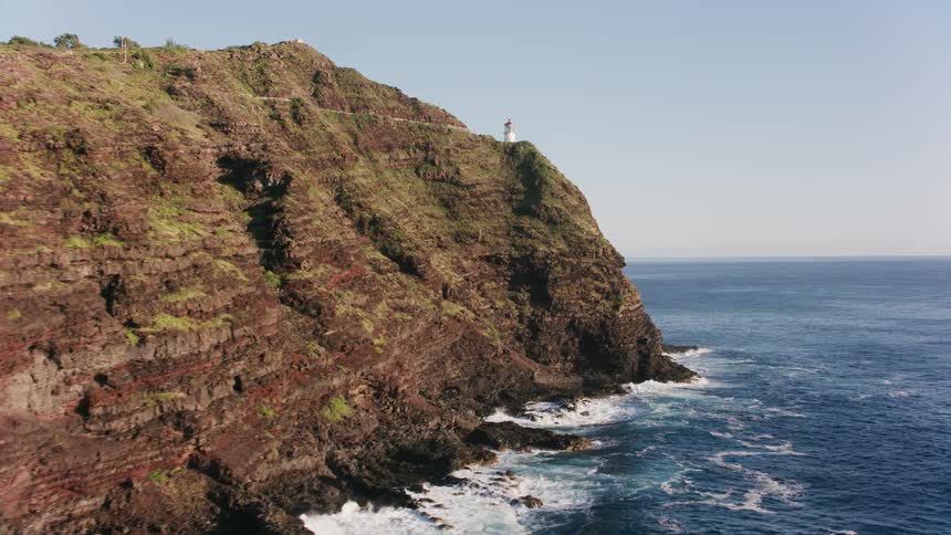 An aerial of a mountain range along a coast with a small lighthouse on the cliff