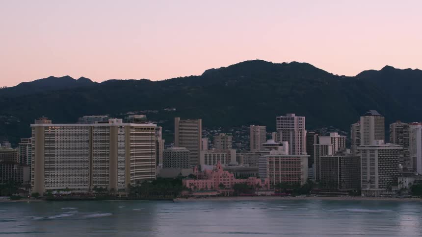 Waves roll into shore and over the beaches along the coast lined with tall buildings and towers of a big city with houses and buildings along the mountainside