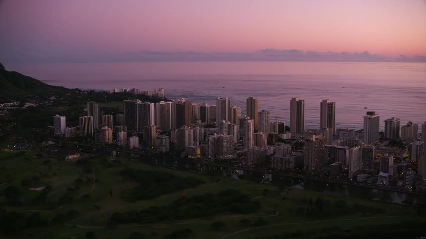 Buildings and towers in a city along the coast stand tall under a pink sky as the sun is setting and small boats sail in the water