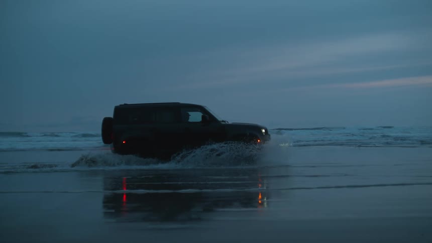 Black land rover defender drives along the beach at dusk