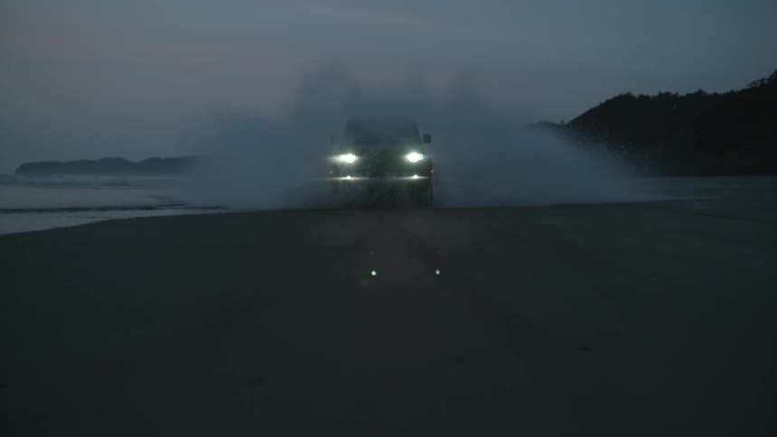 Headlights shine as a truck drives through water on the beach as waves roll into shore with mountains in the distance