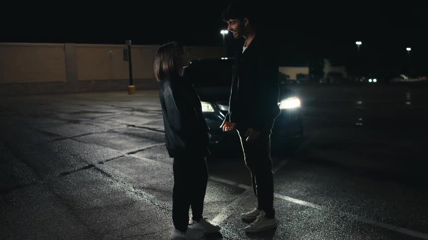 Car headlights shine on a man and a woman standing in a parking lot talking