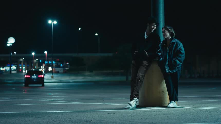 Man with beard jacket pants and white sneakers and woman with leather jacket black pants and white shoes talk with each other at parking lot at night