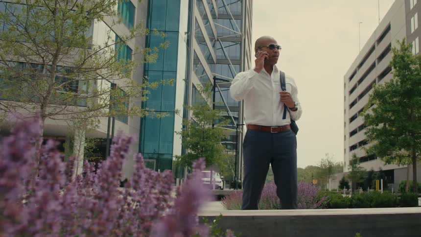 Businessman with sunglasses white shirt brown belt blue pants and backpack talks on smartphone near purple flowers
