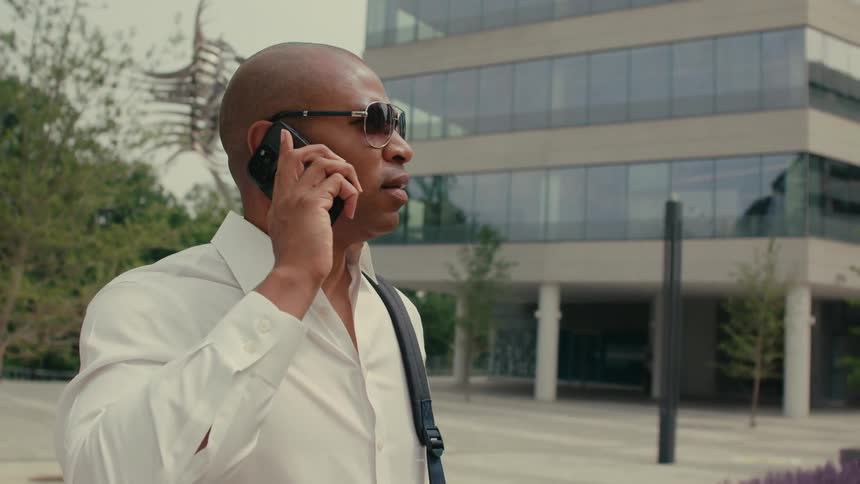A man wearing sunglasses is standing on a city street holding his phone and looking around