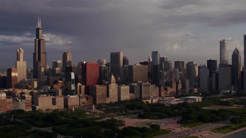 Towers and skyscrapers overlook traffic as it moves on the streets in a metropolitan city along the coast and around the park under a cloudy sky