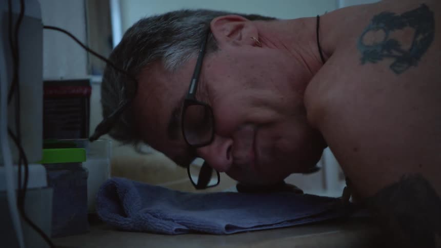 Older with tattoos turns on electric shaver with his chin and bends down to rub his face against it