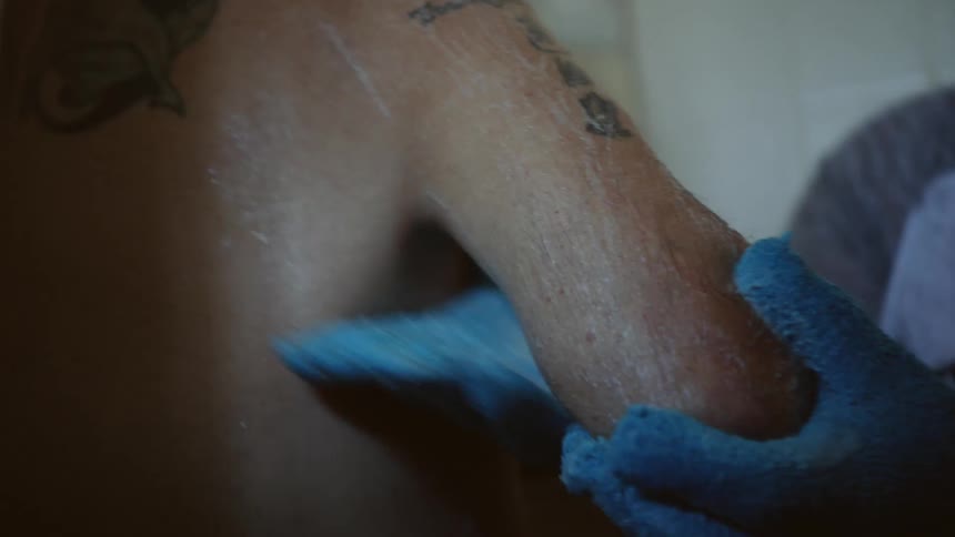 A handicapped man with tattoos on his arm and back is washed with soap by a pair of gloved hands