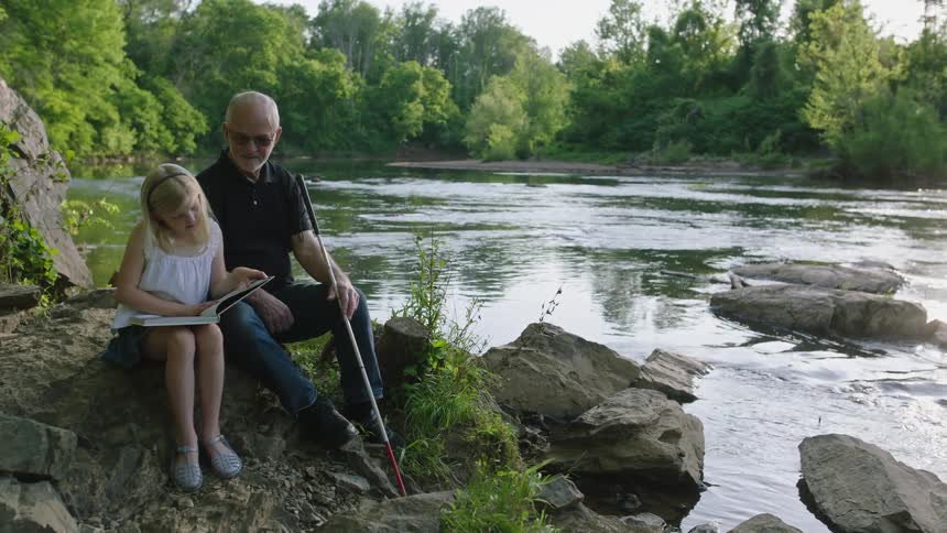 Granddaughter and blind grandfather share a sweet moment reading together by the river