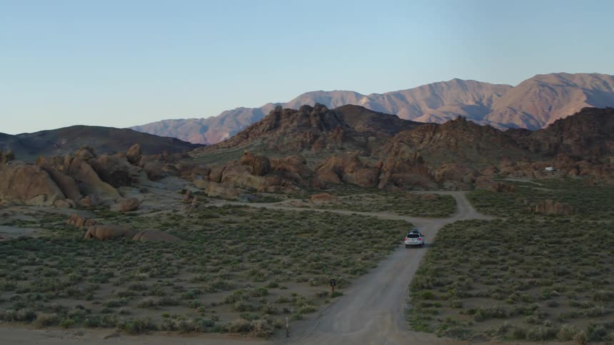 A car drives down a dirt road on a grassy plain surrounded by rock formations until it comes to a crossroads
