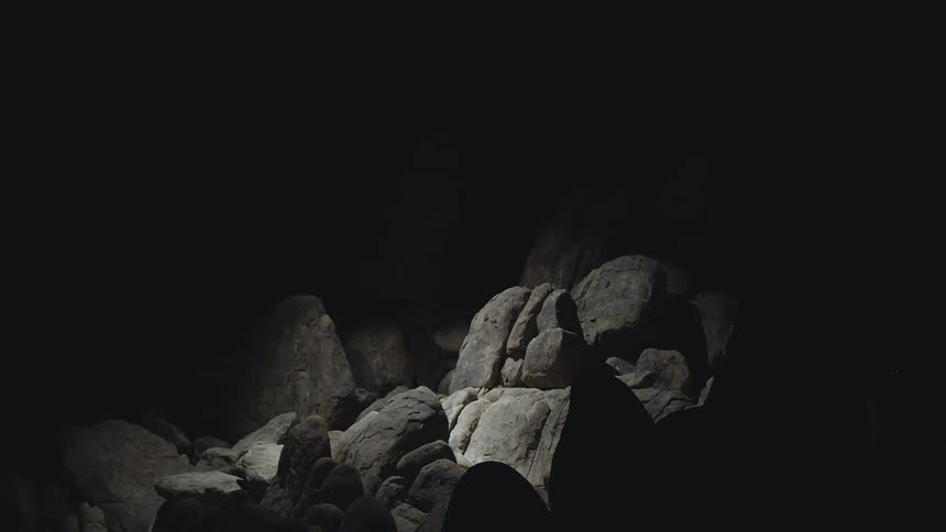 A formation of smooth gray boulders and rocks against a black background and in shadow with a spotlight moving across search light helicopter
