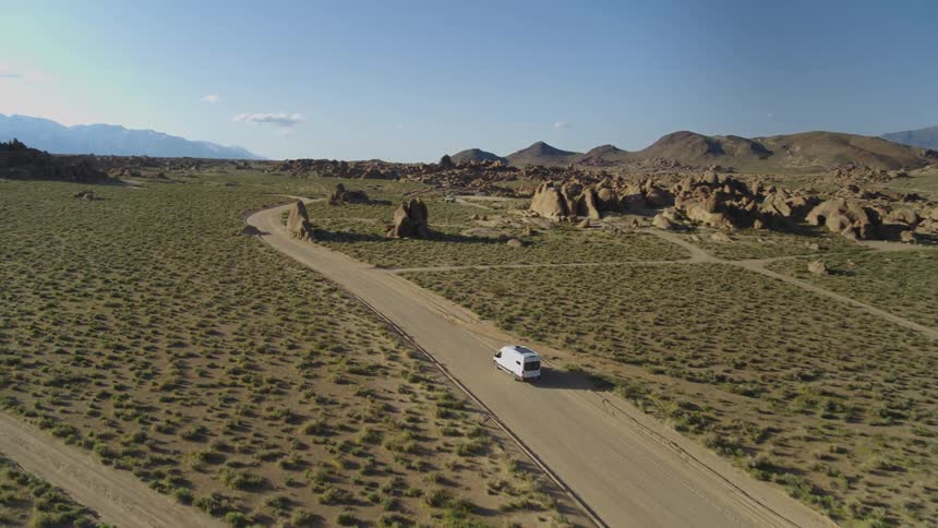 A camper van / delivery truck drives through a dirt road crossing a grassy plain surrounded by rock formations