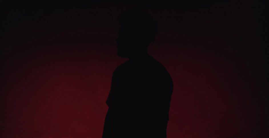 Young man standing against a red background in black silhouette a white spotlight fades up on his shoulder and face serious expression