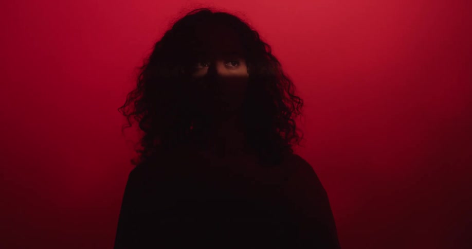 Silhouette of a woman standing with red light in the background and a light exposes her eyes as her hair moves in the breeze