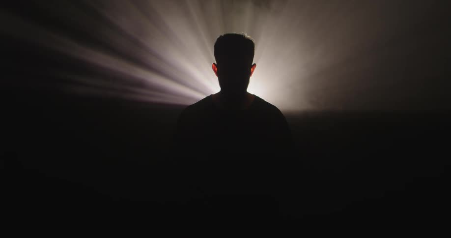 Silhouette of a man standing in the darkness as smoke moves in the rays of light shining behind him