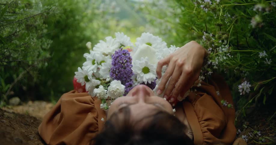A woman is lying on the ground outside with a bouquet of flowers on her chest as she moves her hand over flowers and talks to herself