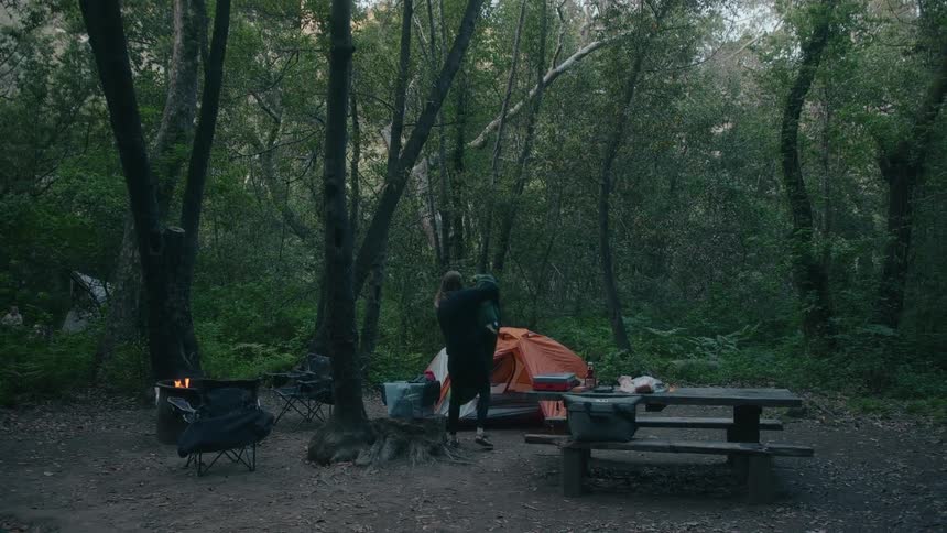 A woman in a forest takes off a blanked off her back while walking towards a tent past a picnic table