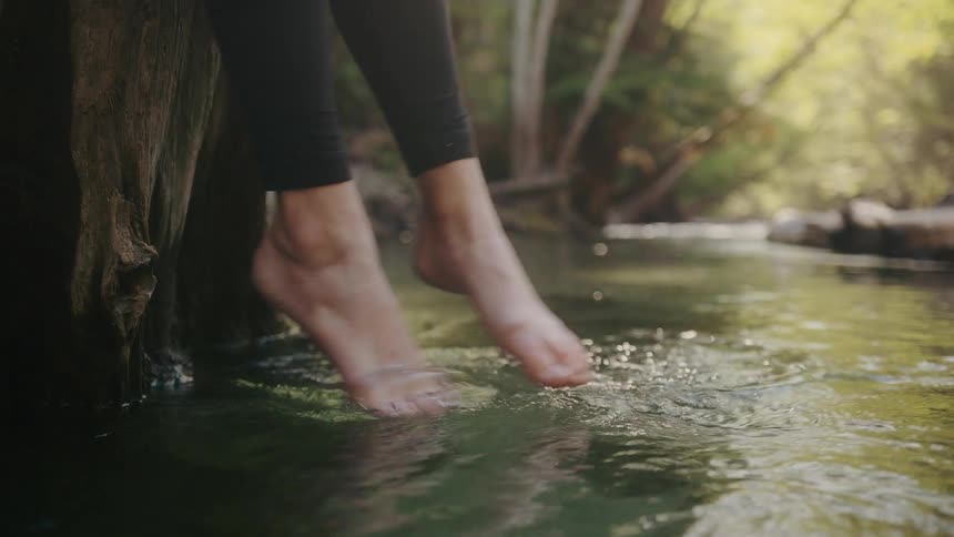 Woman with black leggings sits on tree stump and dips feet in river
