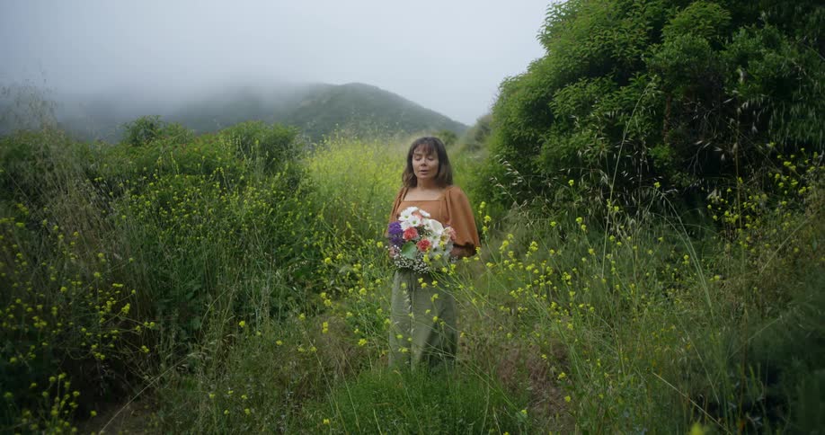 Woman with brown hair blouse and yellow pants holds a bunch of flowers in the green field
