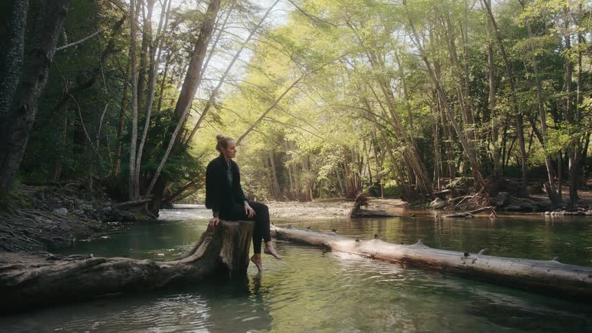 A woman is sitting on a tree stump at the edge of a creek in the woods as she relaxes and dips her toes in the water