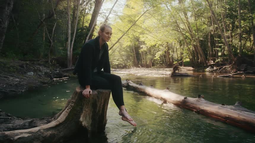 Blonde woman with black jacket and pants sits on tree stump in the river in the forest