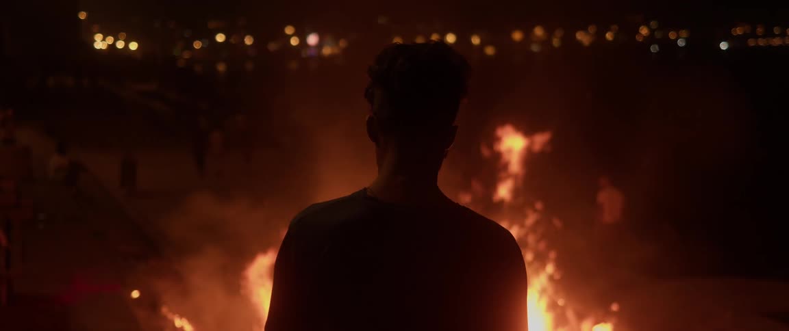 A man stands in front of a tall fire as people dance around him