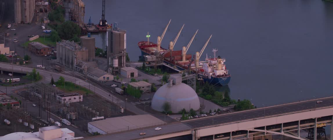 Drone view of a ship docked at port on a waterway as traffic goes by on a nearby bridge going over the river