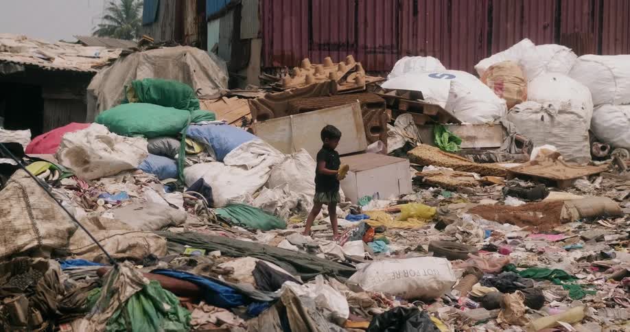 A small boy standing amongst a huge pile of garbage strewn all over the ground as he holds a shoe in his hand
