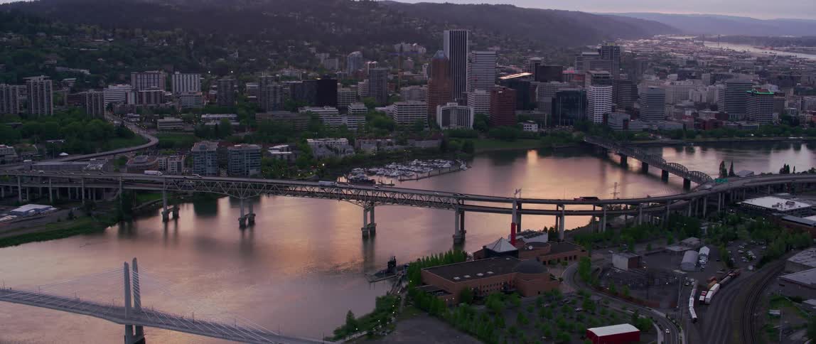 Bridges cross red tinted river reflecting sunlight near downtown with misty mountains in the background