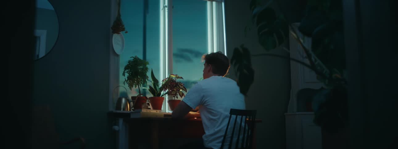 A young man sits in a chair while looking out the window of his home