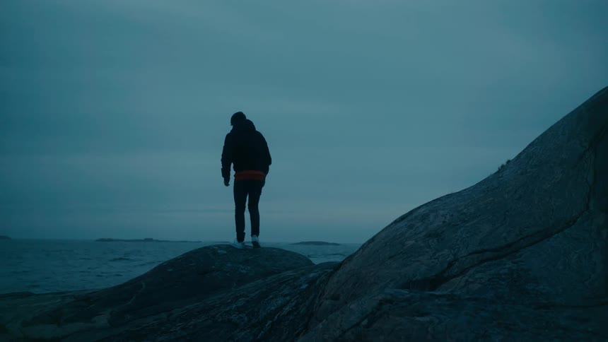 A man is walking on rocks along the coast as he stands looking at the water and waves rolling in to shore