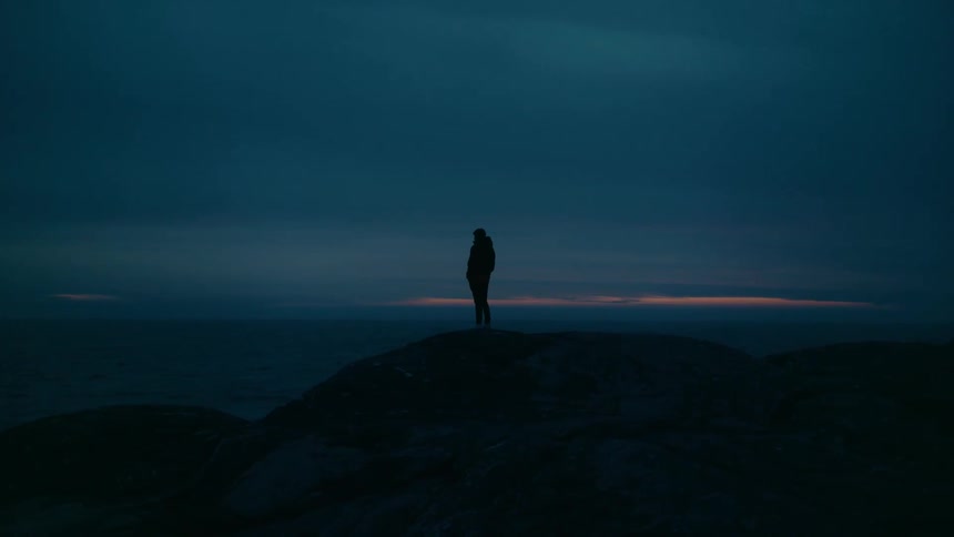 Person stands on shore overlooking ocean and red tinted horizon under an overcast sky