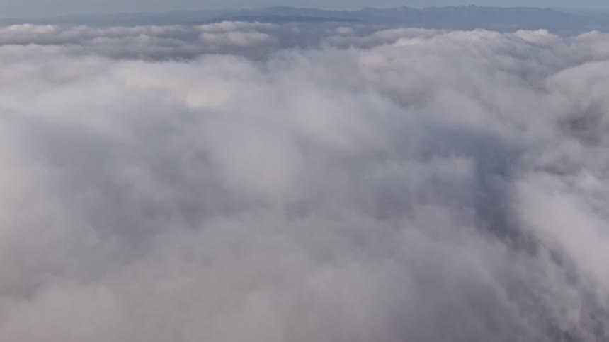 A breathtaking aerial view of the dense marine layer over Los Angeles with a break in the clouds revealing a gorgeous neighborhood with houses close together and mountains on the horizon