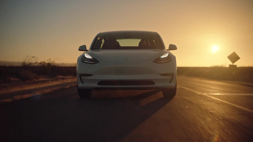 A white automobile travels on a road through the desert as the setting sun shines brightly in the distance