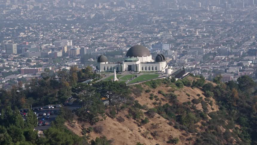 An aerial 4K footage flying low over palm trees to approach and flyby the iconic Griffith Observatory in Los Angeles California on a hazy day showcasing the popular landmark and scientific facilities in Southern California