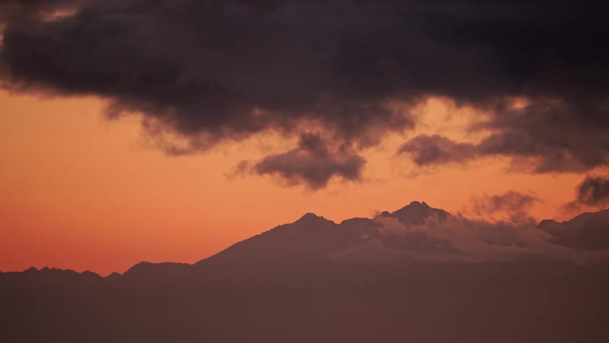 Dark clouds passing in front of a mountain range in front of an orange sky at dawn