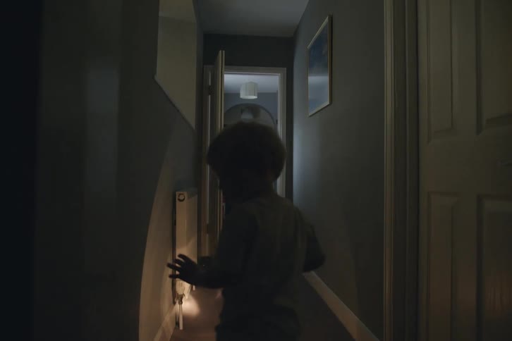 A small child walks down a dark hallway with a flashlight