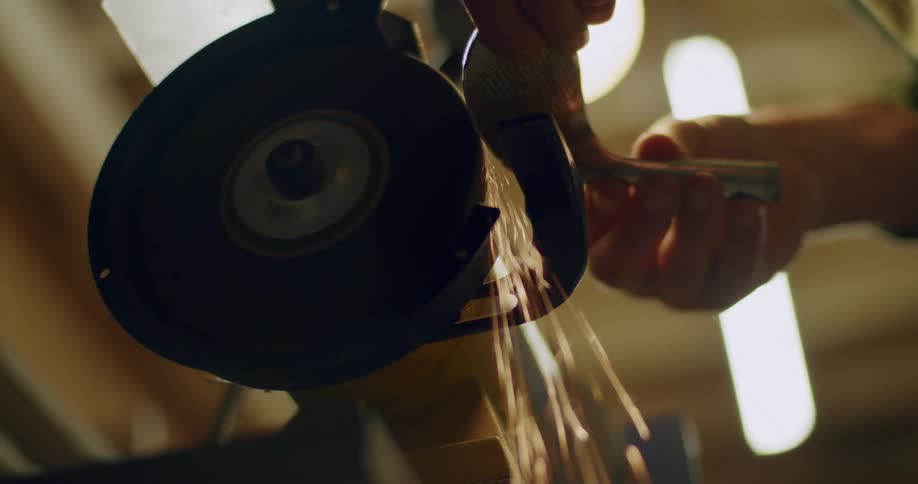 A mans hands are shown holding a metal object and rounding it against a grinding wheel
