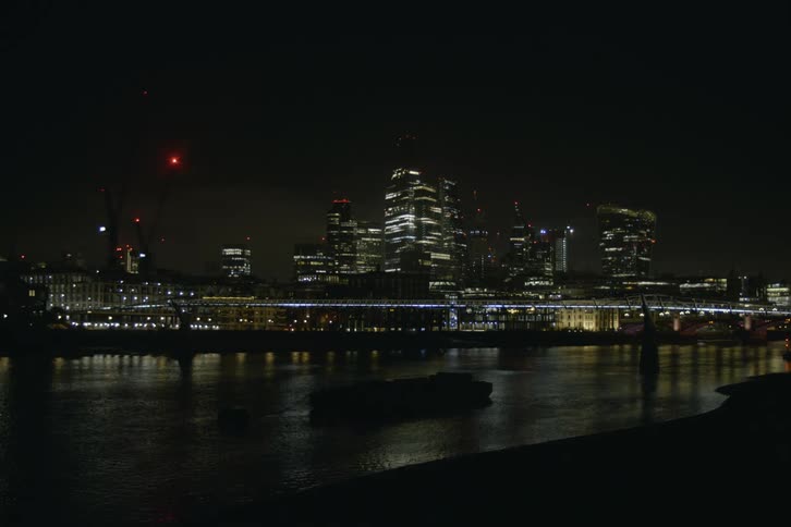 Water lapping onto shore reflects city buildings near bridge at night