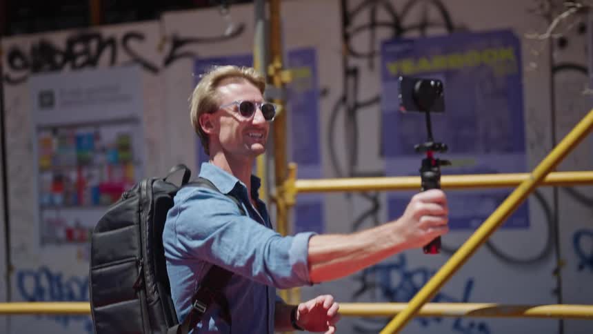 Lively video clip a man is seen holding his phone with a selfie stick while wearing a backpack and glasses he appears to be engaged in a cheerful conversation with someone on his phone all while walking with a sense of enthusiasm