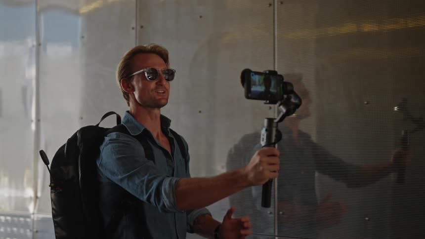 Professional videographers with sunglasses backpack and blue shirt films himself holding a gimbal with smartphone