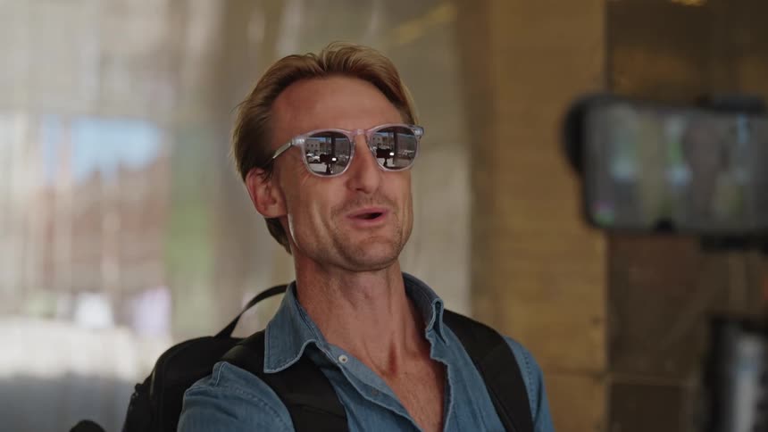 Man with sunglasses blue shirt and backpack films himself on smartphone talking and laughing