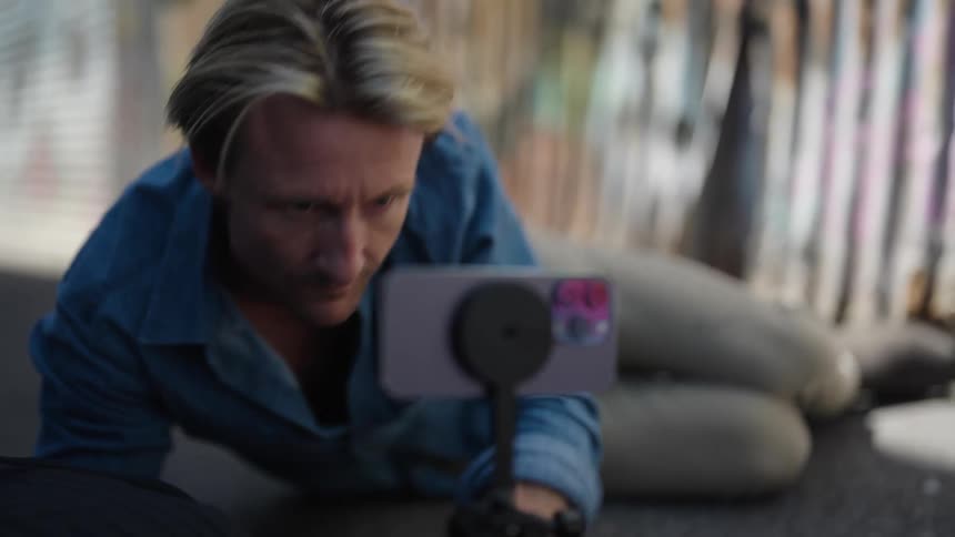 A man lying on the floor on his side adjusting a phone stand so he can use the smart phone as a camera and smiling as he finds the right position