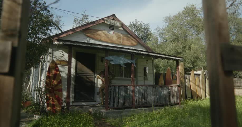 A run down house with a fake shark hanging from a porch and several surf boards leaning into it