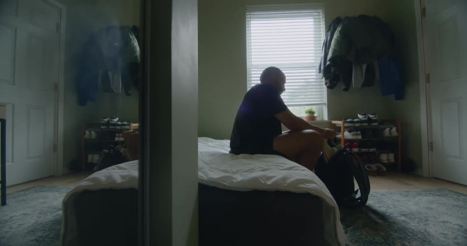 A man packs shoes into a backpack while sitting on a bed during the day