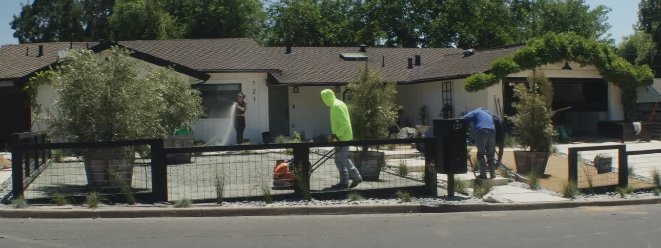 Woman waters yard while work crew works on landscaping of house front yard