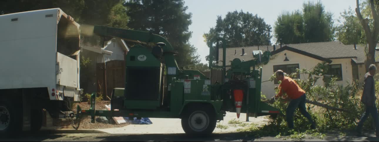 Work crew feeds pruned tree branch into wood chipper parked on street in front of house