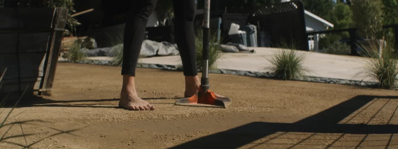 Woman walks barefoot across ground in yard tamping earth with tamping iron