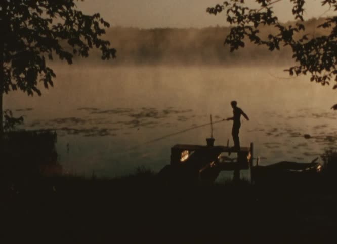 Youth silhouetted against foggy pond catches line and places fishing pole down on dock